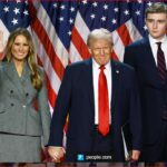 Melania Trump waves while Donald Trump and his son Barron stand beside her in front of a wall of American flags.
