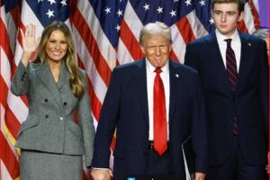 Melania Trump waves while Donald Trump and his son Barron stand beside her in front of a wall of American flags.