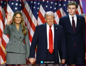 Melania Trump waves while Donald Trump and his son Barron stand beside her in front of a wall of American flags.