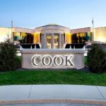 Exterior view of a modern corporate building with a lit fountain and a large illuminated COOK sign in front, surrounded by green lawns and shrubs at dusk.