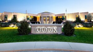 Exterior view of a modern corporate building with a lit fountain and a large illuminated COOK sign in front, surrounded by green lawns and shrubs at dusk.