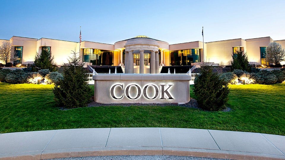 Exterior view of a modern corporate building with a lit fountain and a large illuminated COOK sign in front, surrounded by green lawns and shrubs at dusk.