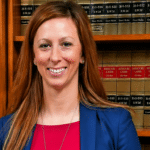 Smiling woman in a blue blazer and red top stands in a law library with shelves of case books behind her; USA Herald logo at the bottom right.