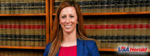 Smiling woman in a blue blazer and red top stands in a law library with shelves of case books behind her; USA Herald logo at the bottom right.