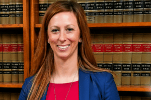Smiling woman in a blue blazer and red top stands in a law library with shelves of case books behind her; USA Herald logo at the bottom right.