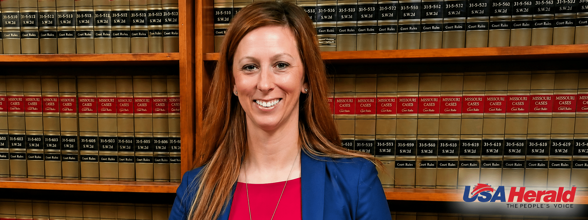Smiling woman in a blue blazer and red top stands in a law library with shelves of case books behind her; USA Herald logo at the bottom right.