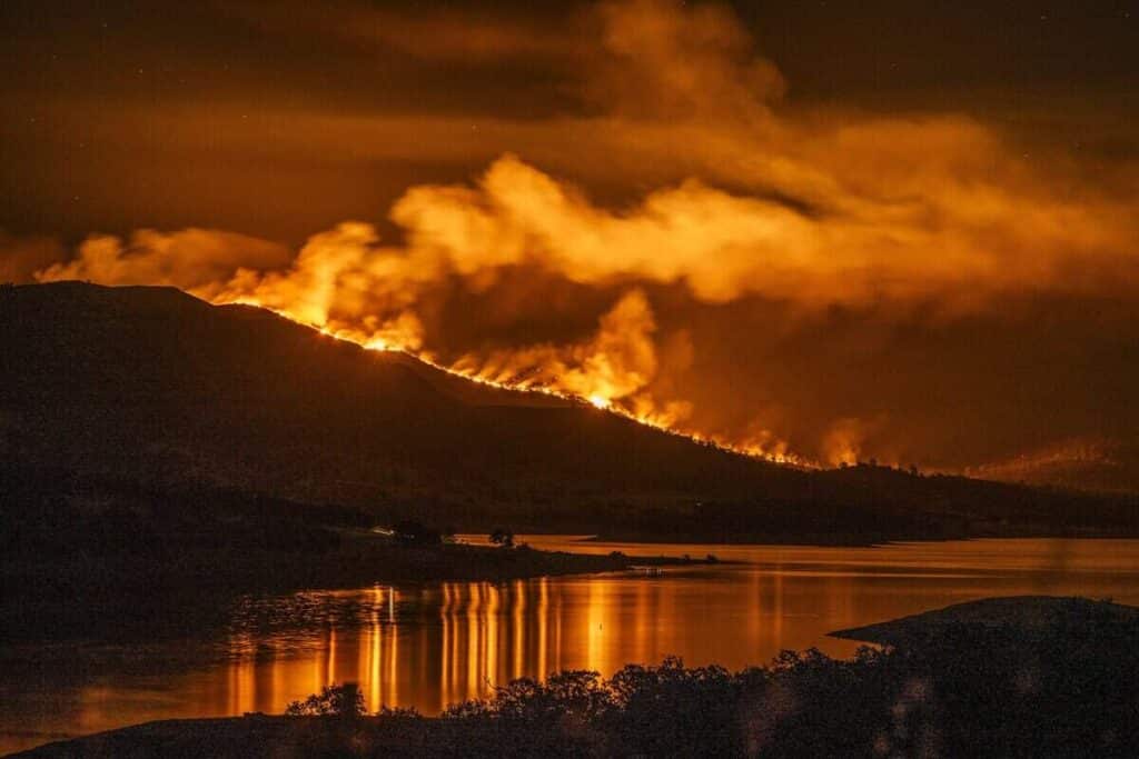 Nighttime wildfire on a hillside with flames and smoke, reflecting orange light on a calm river