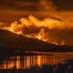 Nighttime wildfire on a hillside with flames and smoke, reflecting orange light on a calm river