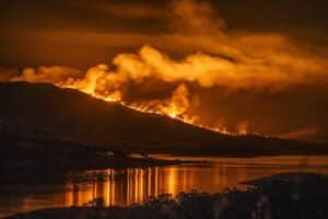 Nighttime wildfire on a hillside with flames and smoke, reflecting orange light on a calm river