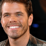 Close-up portrait of a smiling man in a plaid shirt, dark background, with the USA Herald logo in the lower right corner.
