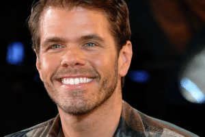 Close-up portrait of a smiling man in a plaid shirt, dark background, with the USA Herald logo in the lower right corner.