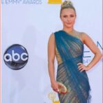 Woman posing on the Emmy Awards red carpet in a blue and gold one-shoulder gown, holding a striped clutch.