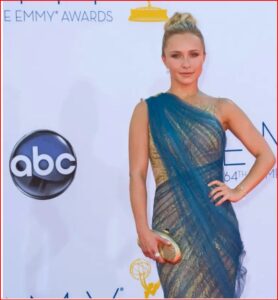 Woman posing on the Emmy Awards red carpet in a blue and gold one-shoulder gown, holding a striped clutch.
