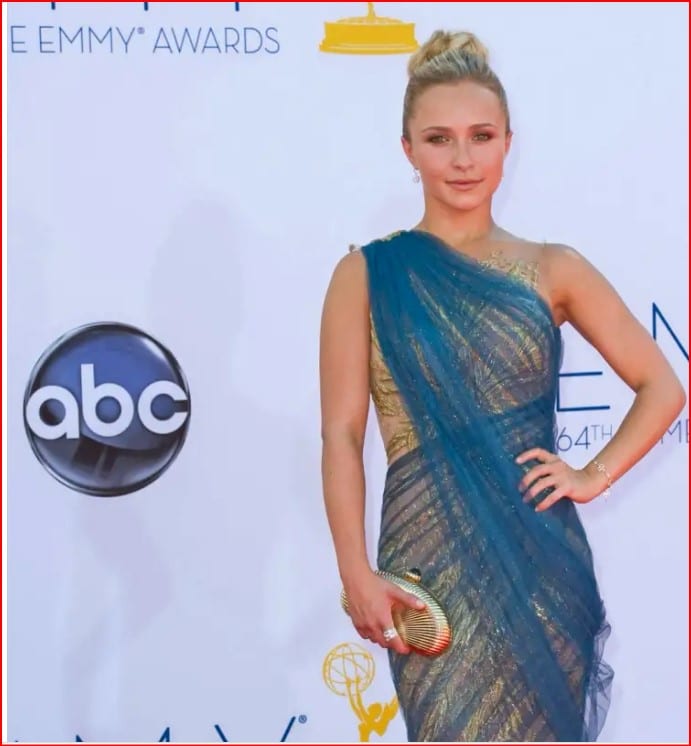 Woman posing on the Emmy Awards red carpet in a blue and gold one-shoulder gown, holding a striped clutch.