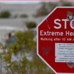 Red stop sign with text 'STOP Extreme Heat Danger' and 'Walking after 10 AM not recommended'; blurred figures walking in a desert landscape in the background.