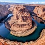 Aerial view of red canyon walls with a blue-green river looping around a central sandstone promontory in a horseshoe bend.