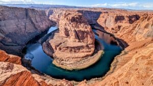Aerial view of red canyon walls with a blue-green river looping around a central sandstone promontory in a horseshoe bend.