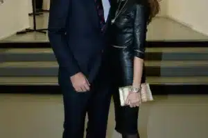 Couple posing together at an indoor event; man in a navy suit, woman in a black leather dress with a gold clutching purse in hand?