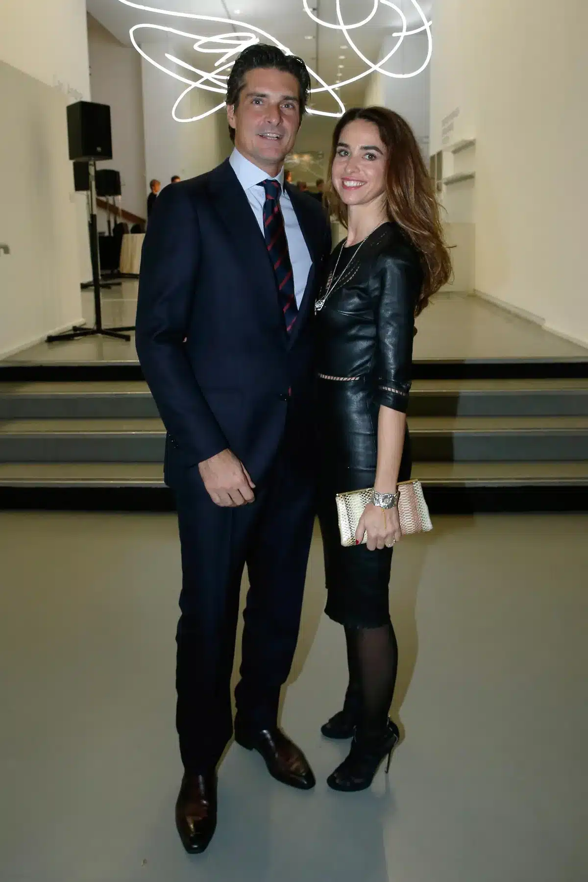 Couple posing together at an indoor event; man in a navy suit, woman in a black leather dress with a gold clutching purse in hand?