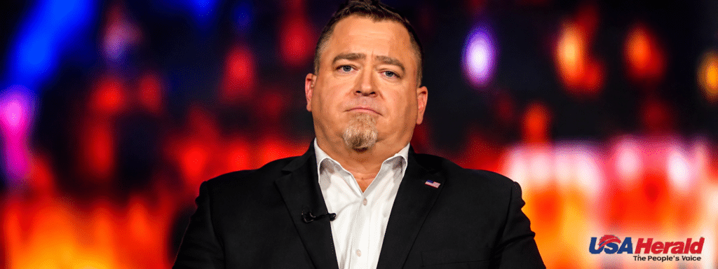 Man in a black suit and white shirt on stage, with a lapel pin, speaking or presenting under colorful lights; USA Herald logo visible bottom right