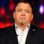 Man in a black suit and white shirt on stage, with a lapel pin, speaking or presenting under colorful lights; USA Herald logo visible bottom right