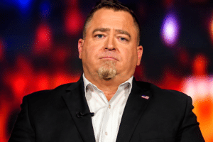 Man in a black suit and white shirt on stage, with a lapel pin, speaking or presenting under colorful lights; USA Herald logo visible bottom right