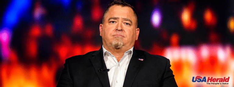 Man in a black suit and white shirt on stage, with a lapel pin, speaking or presenting under colorful lights; USA Herald logo visible bottom right