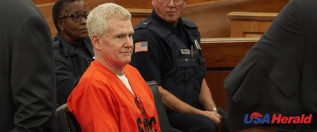 Man in an orange inmate jumpsuit sits in a courtroom beside uniformed officers; wooden benches visible behind him, USA Herald logo bottom right.