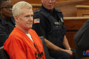Man in an orange inmate jumpsuit sits in a courtroom beside uniformed officers; wooden benches visible behind him, USA Herald logo bottom right.