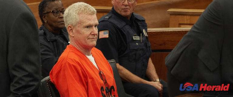 Man in an orange inmate jumpsuit sits in a courtroom beside uniformed officers; wooden benches visible behind him, USA Herald logo bottom right.