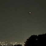 Two bright lights visible in a dark night sky above a city skyline, with tree silhouettes in the foreground and a USA Herald watermark in the bottom-right corner.