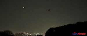 Two bright lights visible in a dark night sky above a city skyline, with tree silhouettes in the foreground and a USA Herald watermark in the bottom-right corner.