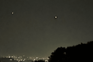 Two bright lights visible in a dark night sky above a city skyline, with tree silhouettes in the foreground and a USA Herald watermark in the bottom-right corner.