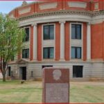Red-brick, neoclassical campus building with white columns and a curved façade, on a grassy quad with a granite memorial in front.