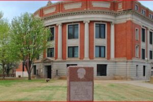 Red-brick, neoclassical campus building with white columns and a curved façade, on a grassy quad with a granite memorial in front.
