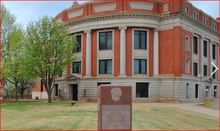Red-brick, neoclassical campus building with white columns and a curved façade, on a grassy quad with a granite memorial in front.