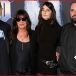 Four people pose together on a red carpet at a formal event, including a bearded man in a cap, a woman in sunglasses, a younger woman in a black dress, and another bearded man in a black jacket holding a paper.
