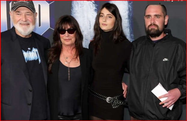 Four people pose together on a red carpet at a formal event, including a bearded man in a cap, a woman in sunglasses, a younger woman in a black dress, and another bearded man in a black jacket holding a paper.