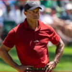 Man in a red polo shirt and black cap stands with hands on hips on a golf course, crowd blurred in the background.