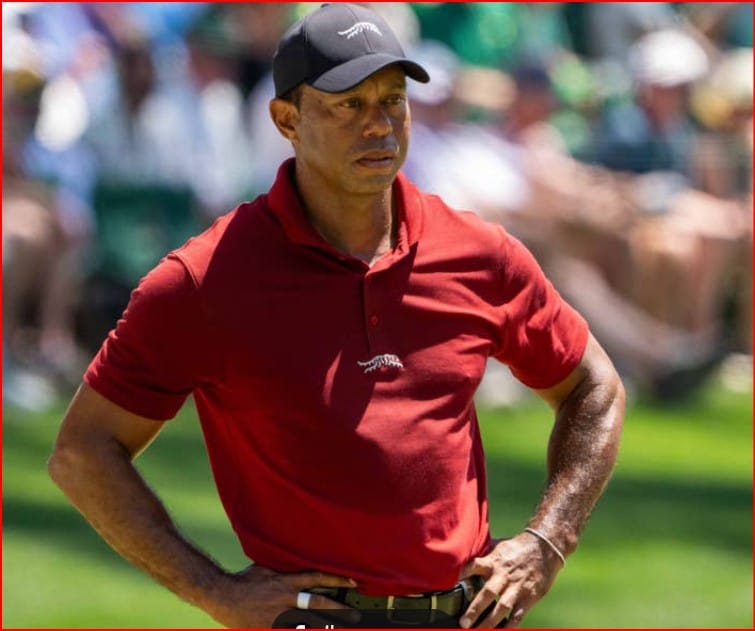 Man in a red polo shirt and black cap stands with hands on hips on a golf course, crowd blurred in the background.