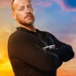 Man with arms crossed posing against a vibrant sunset sky, wearing a black long-sleeve shirt.