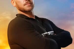Man with arms crossed posing against a vibrant sunset sky, wearing a black long-sleeve shirt.