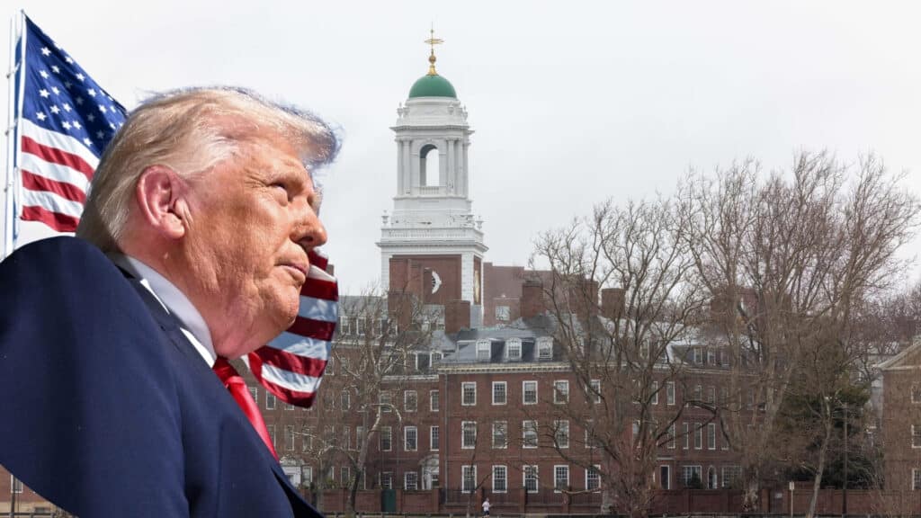Man in a dark suit and red tie seen in profile, with an American flag behind him and a brick campus building with a green-domed tower in the background.