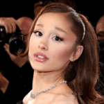 Woman wearing a diamond necklace posing on a red carpet, photographers in the background.