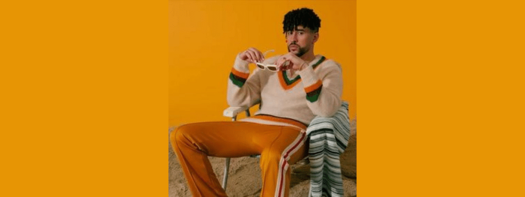 Man wearing a cream sweater with orange and green stripes sits in a chair, holding white sunglasses against an orange backdrop.