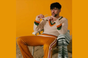 Man wearing a cream sweater with orange and green stripes sits in a chair, holding white sunglasses against an orange backdrop.