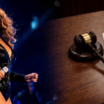 Female singer performing on stage, holding a microphone, with a judge's gavel and legal documents visible on a desk to the right.