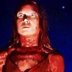 Woman with red lighting on face and neck, wide-eyed expression, looking upward against a dark blue, speckled background.
