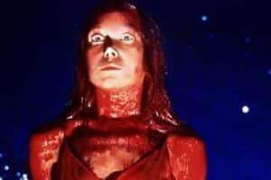 Woman with red lighting on face and neck, wide-eyed expression, looking upward against a dark blue, speckled background.