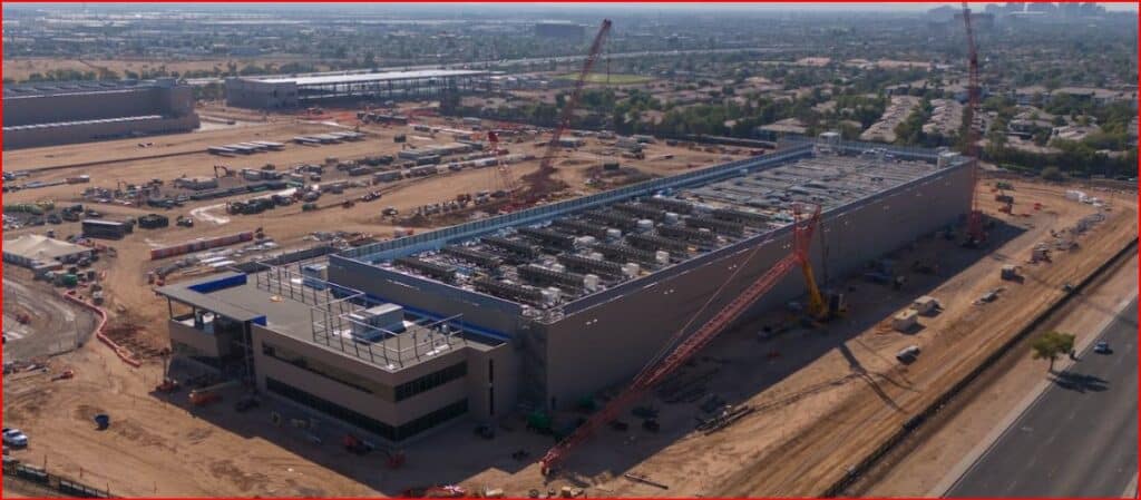 A long industrial building under construction seen from above, with multiple cranes and a dusty work site surrounding the structure.